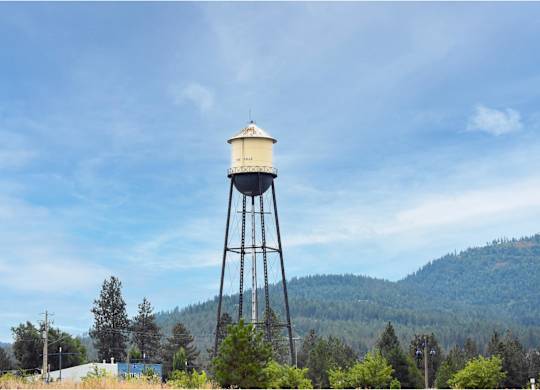 A water tower stands tall in a field with trees and hills in the background.