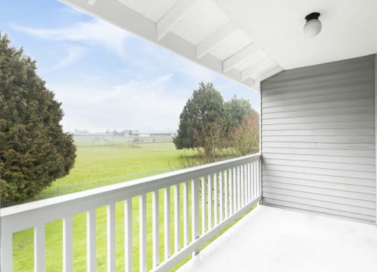 A private, covered apartment balcony with a white railing looks out to grass fields and trees.at The Trail, Snohomish, 98290