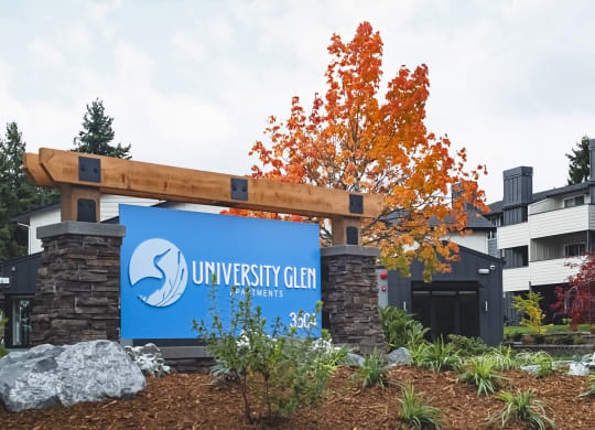 Community entrance with blue 'University Glen' sign surrounded by green plants, bushes, large rocks, and trees at University Glen, University Place, WA