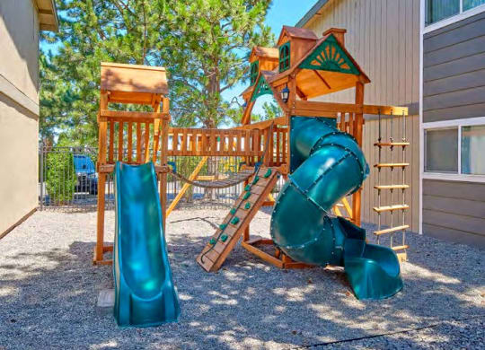 A playground with two slides, a swing set, and a small rock climbing wall surrounded by pea gravel and trees in the background.at Wildflower, Washington