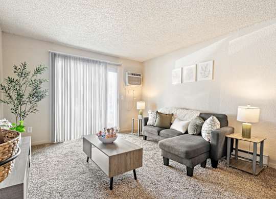 A living room with a grey couch, a coffee table, and a plant A row of apartment buildings with green lawns in front A bedroom with a bed, nightstand, and window at Wildflower, Kennewick, WA 99336.