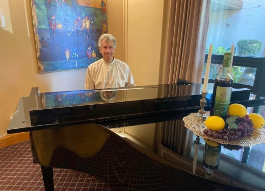 a man playing a piano in a room with a table with fruit on it at River Commons Senior Living, Redding California