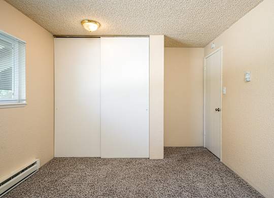 A room with a carpeted floor, a window with blinds, a white door, and a light fixture on the ceiling.