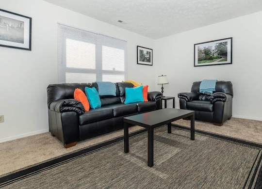 A black leather couch with orange and blue pillows sits in a living room with a coffee table in front of it.