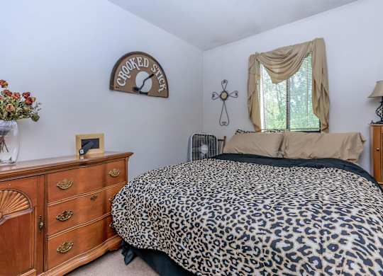 A leopard print bed sits in a bedroom with a wooden dresser and a cross hanging on the wall.