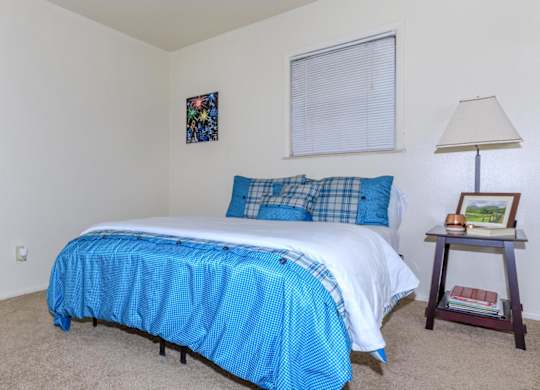 A bed with a blue and white comforter is in a room with a lamp and picture frame on a side table.