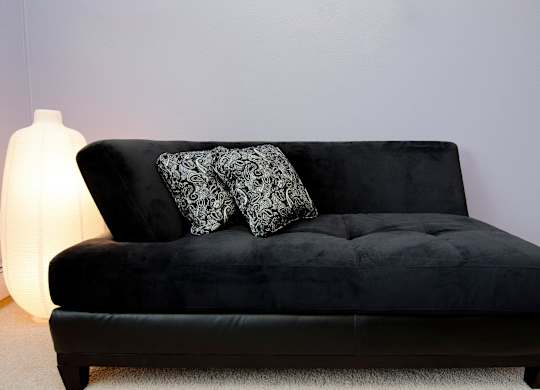 A black couch with two patterned pillows sits in a room with a white lamp.