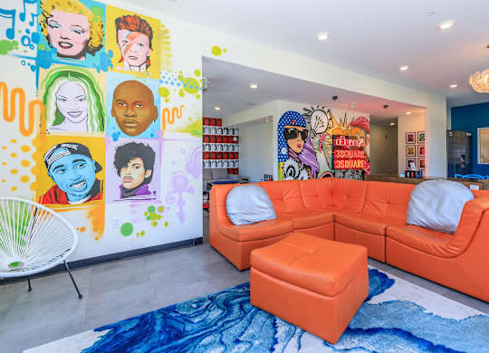 A brightly colored living room with a large orange sofa and a mural of various faces on the wall.