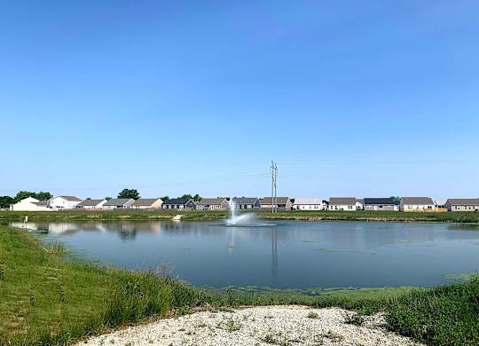 A serene lake with a fountain in the middle, surrounded by houses and a clear blue sky.