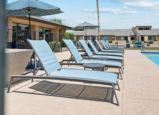 A row of sun loungers are on a concrete patio next to a pool.