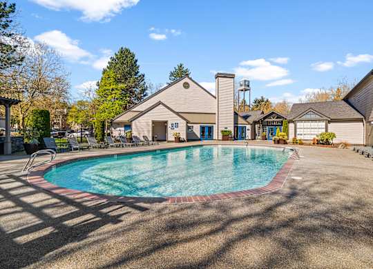 A large outdoor swimming pool surrounded by a patio area with chairs and a building in the background.