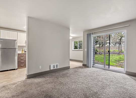 A spacious, empty room with a grey carpet and a sliding glass door leading to a green outdoor area.