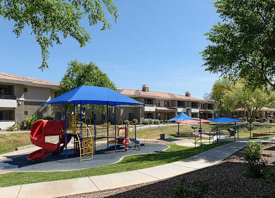 A playground with a red slide and blue umbrellas is surrounded by apartment buildings.