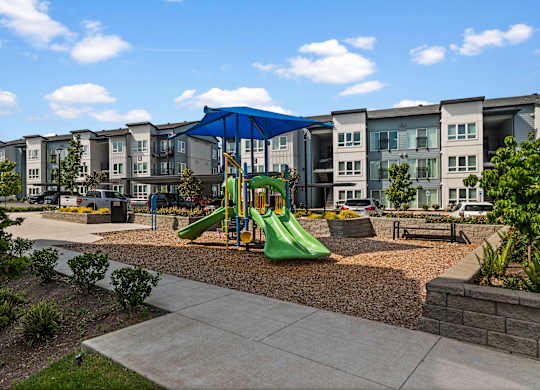 A playground with a green slide and a blue canopy is in front of apartment buildings.