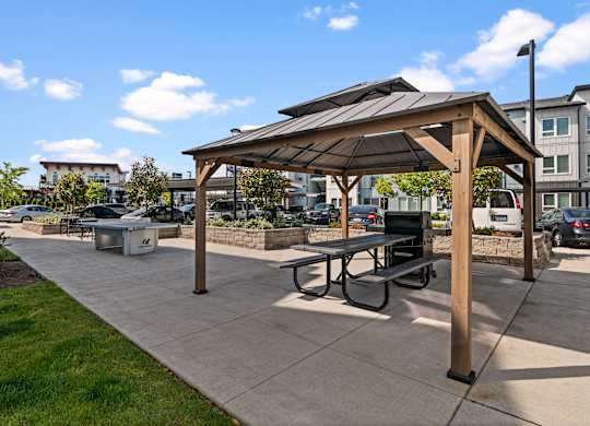 A wooden pavilion with a black roof is surrounded by benches and a concrete walkway.