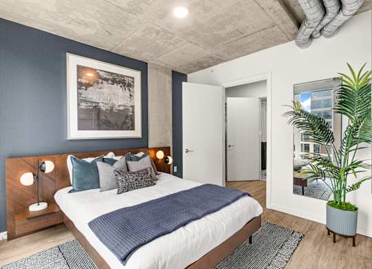 A bedroom with a bed, a nightstand with lamps, a potted plant, and a framed picture on the wall.