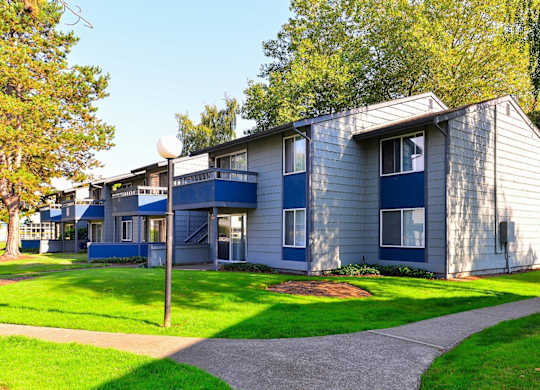 A small building with a grey and blue exterior is surrounded by a grassy area and trees. at The Lakes at Fife, Fife, WA