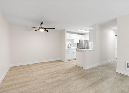 A room with a ceiling fan and light fixture, and a kitchen area in the background.