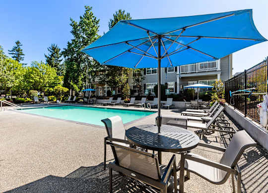 A blue umbrella shades a table and chairs by a pool.