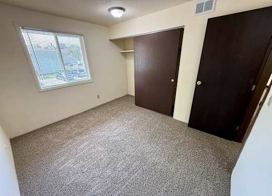 A room with a carpeted floor, a window with blinds, and two brown doors. at Winding Hills Apartments, Iowa