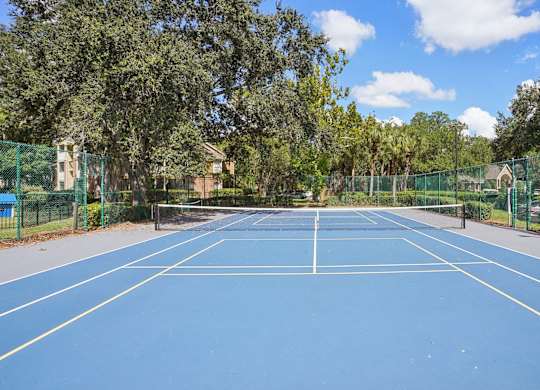 A blue tennis court surrounded by green fencing and trees.