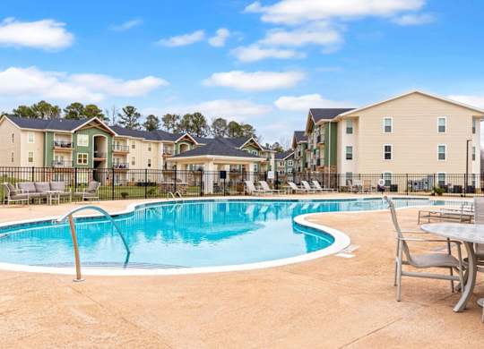 A swimming pool surrounded by chairs and tables in front of apartment buildings.