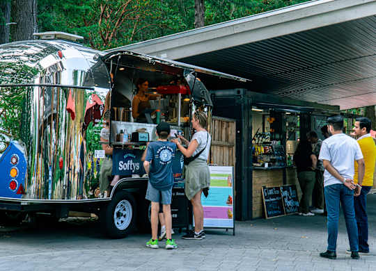 A food truck with a silver exterior and a blue and white logo on the side is parked outside a building with people standing in front of it.