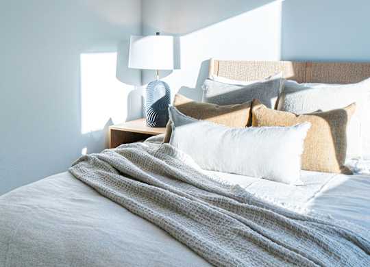 A bed with a grey blanket and pillows is in the foreground with a lamp on a side table in the background.