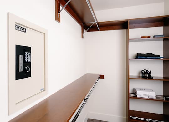 A white safe with a digital keypad is on the left side of a room with a wooden shelf on the right.