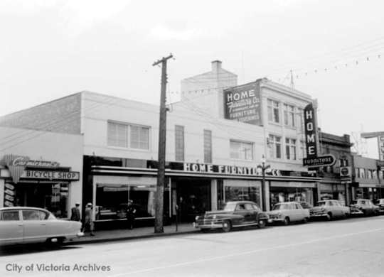A black and white photo of a street with a Home Furnishings store and a Bicycle Shop.
