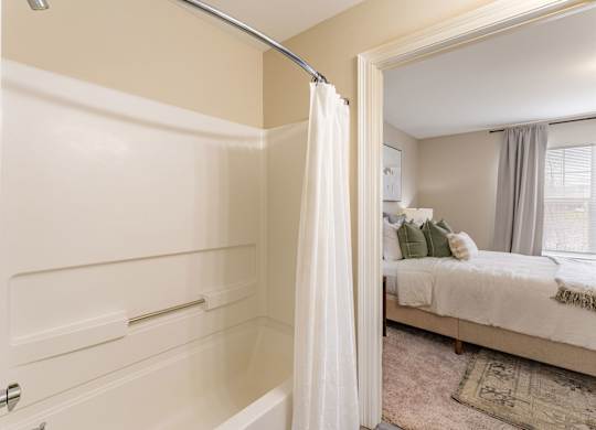 A white bathroom with a shower and a bedroom in the background.