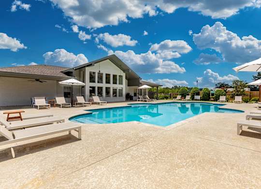 A large swimming pool in front of a house with a patio and lounge chairs.