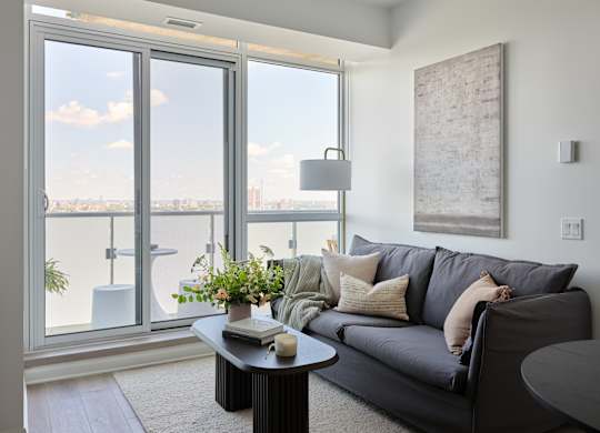 A living room with a grey couch and a large window overlooking the city.
