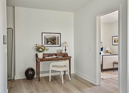 A room with a desk, chair, and vase on the floor.