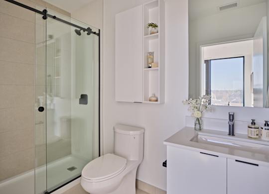 A modern bathroom with a glass shower door, white toilet, and a white sink with a mirror above it.