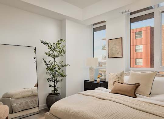 A bedroom with a large bed, a chair, a potted plant, and a mirror.