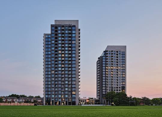Two modern high-rise buildings stand side by side with a grassy field in the foreground.