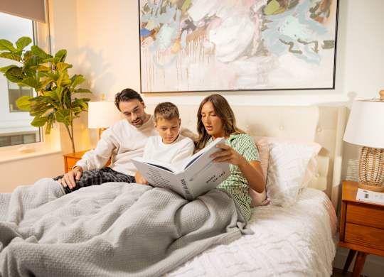 A family of three is reading a book together on a bed.
