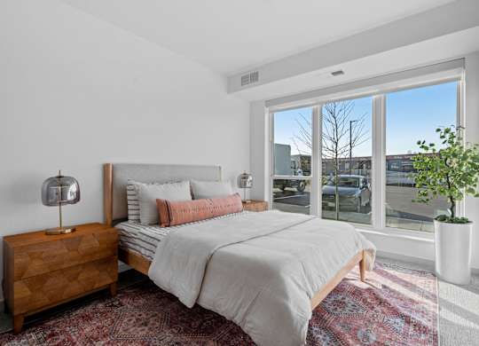 A bedroom with a large bed, a wooden nightstand, and a window overlooking a cityscape.