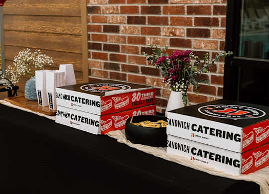 A table with a black cloth and several boxes of Sandwich Catering on it.