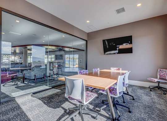 A conference room with a long table and chairs, a television on the wall, and a view of the outside through a glass wall.