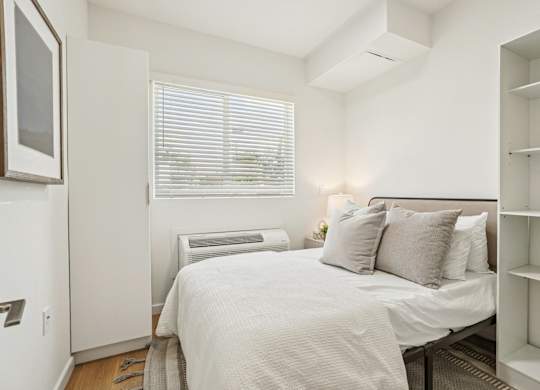 A white bedroom with a bed, pillows, and a window with blinds.