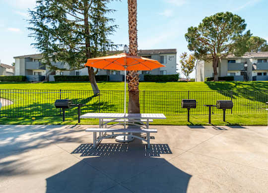 A picnic table with an orange umbrella is in the middle of a concrete area with a tree and mailboxes in the background at Chaparral Apartments, Palmdale, CA, 93551