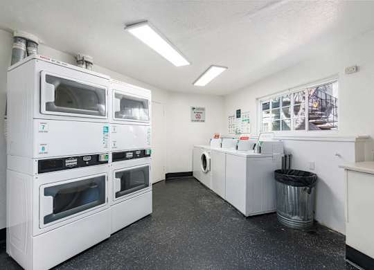 A clean, white-walled commercial kitchen with stainless steel appliances and a trash can.