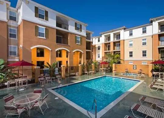A swimming pool surrounded by chairs and umbrellas in front of apartment buildings.