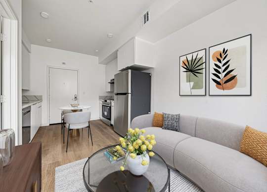 A modern living room with a grey sofa, a glass coffee table, and a dining area in the background.