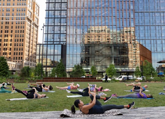 A group of people doing yoga in a park with a glass building in the background.