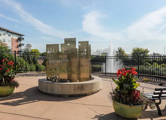 A fountain in the middle of a park with benches and flower pots.