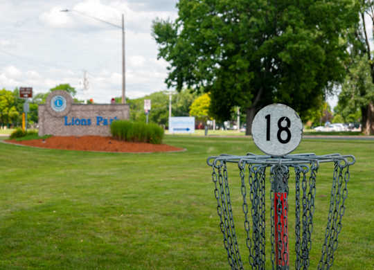 A disc golf basket with the number 18 on it is in the foreground of a grassy area with a sign that says Lions Park in the background.