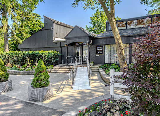 A building with a dark grey exterior and a black roof is surrounded by a well-manicured garden with a variety of plants and trees. at Commons At Waters Edge, Columbus Ohio